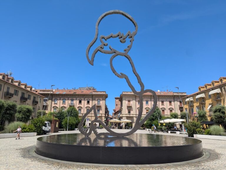 Moderne Skulptur „Alba“ von Valerio Berruti auf der Piazza Michele Ferrero in Alba, Piemont. Das Kunstwerk zeigt die stilisierte Umrissfigur eines knienden Kindes über einem runden Wasserbecken, eingerahmt von klassizistischen Gebäuden und blauem Himmel.