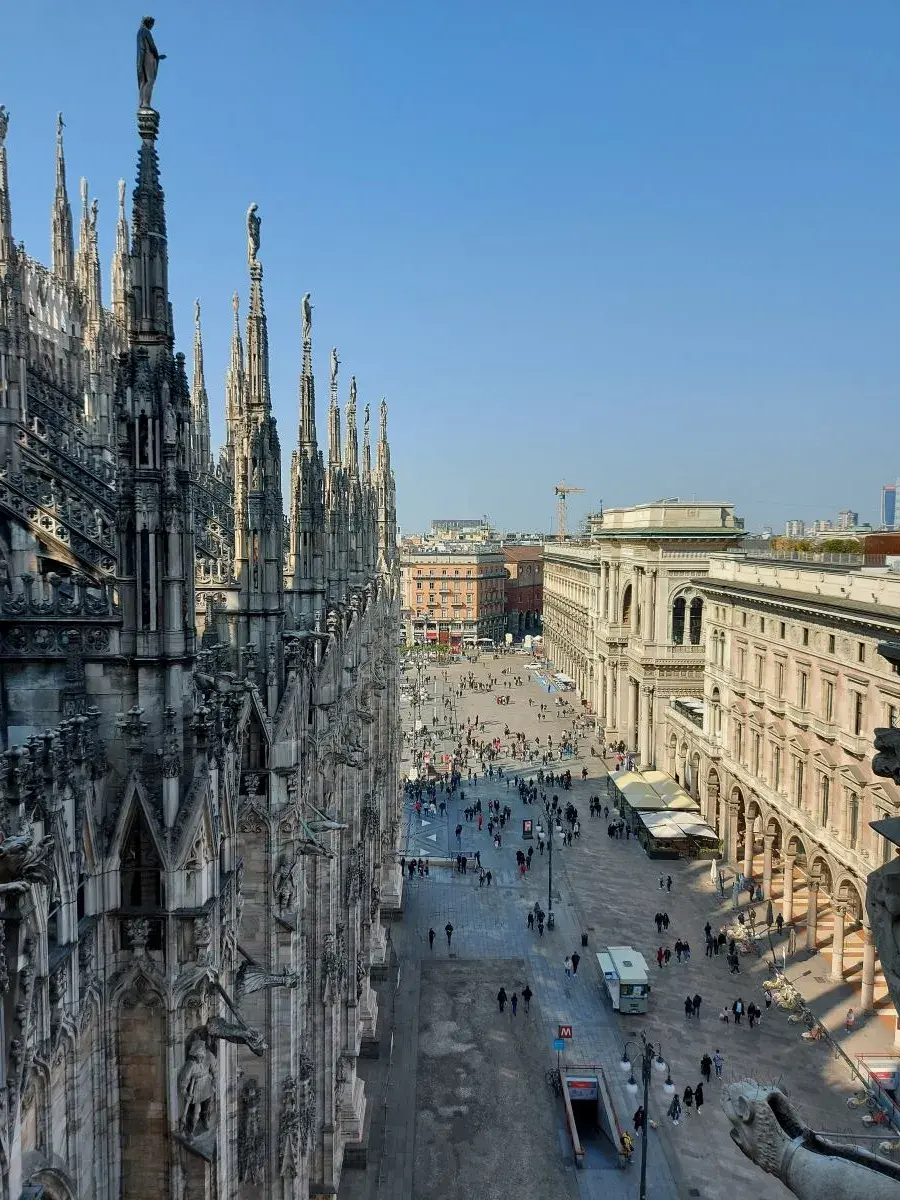 View from the roof of Milan Cathedral to the side street below. Many people walk along medieval palazzi towards the cathedral square.