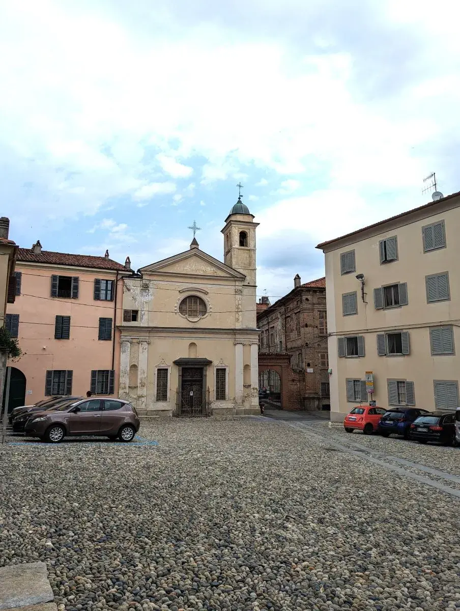 Die gelbe Kirche Chiesa San Nicola da Tolentino von Ivrea steht mittig auf einem großen Platz aus Kopfsteinplaster mit einigen, geparkten Autos davor