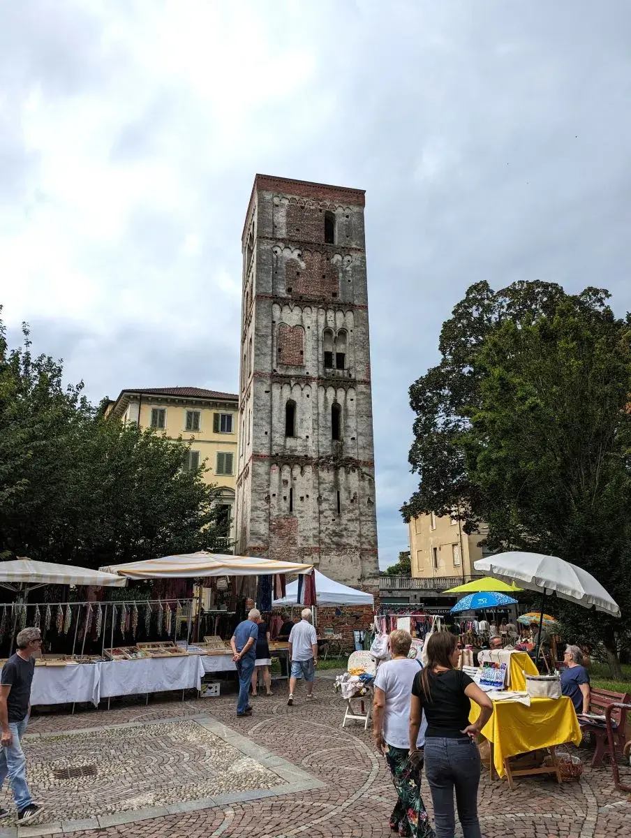Einige Marktstände, mit neugierigen Menschen davor, stehen auf dem Platz unterhalb des Glockenturm Santo Stefano von Ivrea