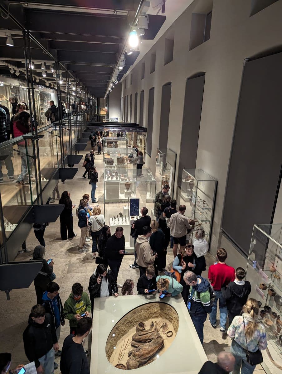 An aerial view of the bustling 4th floor of the Egyptian Museum Turin, showcasing visitors observing various exhibits. The image features a mix of glass cases filled with artifacts, museum-goers in casual attire, and a prominent circular display with ancient pottery at the center.