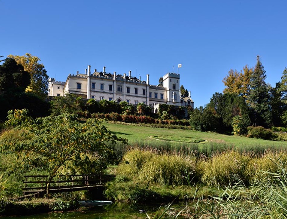 The majestic Castello Dal Pozzo, one of the luxury hotels at Lake Maggiore, surrounded by lush gardens and overlooking a serene landscape. This historic hotel boasts a pristine white façade with multiple balconies and a classical architectural style, set against a backdrop of mature trees and a clear blue sky.