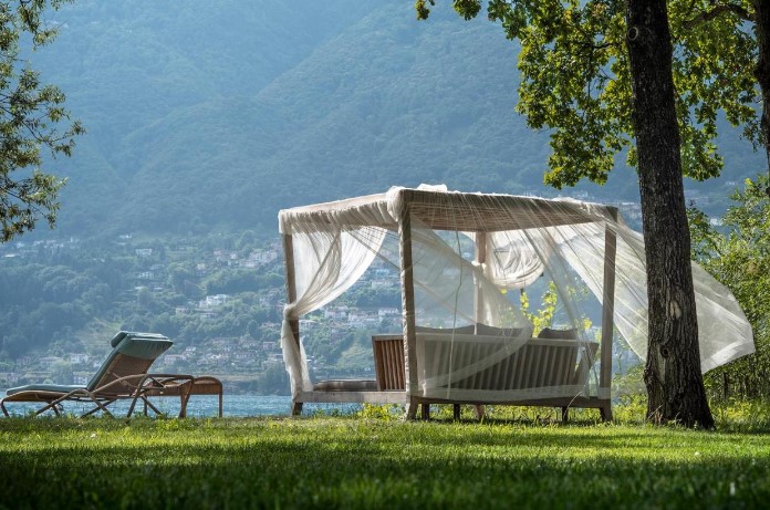 Outdoor relaxation area at Castello del Sole Beach Resort & SPA, featuring a canopy bed and lounge chairs, offering a tranquil retreat at a luxury hotel on Lake Maggiore. The setting includes a pristine lawn with views of distant hills and the shimmering lake, ideal for relaxation and taking in the natural beauty of the area.