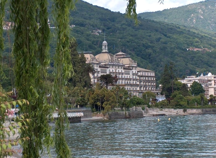 Grand Hotel des Iles Borromées & SPA, a luxury Lake Maggiore hotel , set against a backdrop of mountains and situated by the lakeside, visible through hanging willow branches. The grandeur of this palatial hotel is highlighted by its ornate architecture and the lush greenery surrounding it, offering a peaceful escape with picturesque views.