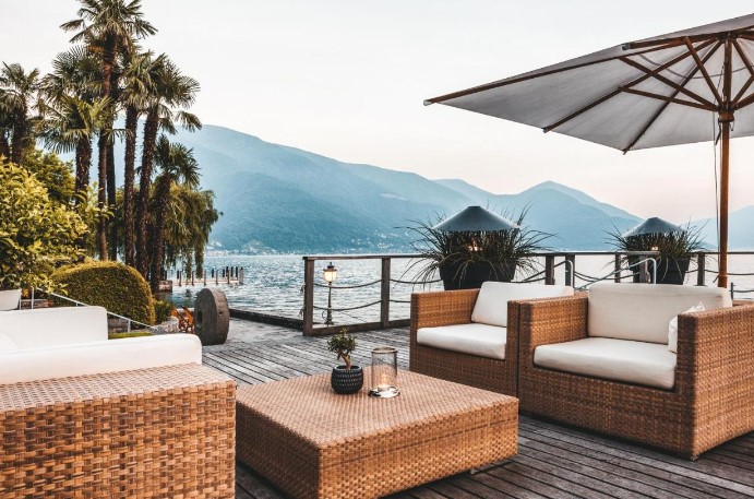 Hotel Eden Roc's lakeside patio setup with modern wicker furniture under a stylish umbrella, showcasing a panoramic view of the mountains, epitomizing luxury at Lake Maggiore. This elegantly arranged outdoor lounge offers guests a chic and comfortable spot to enjoy the stunning lakefront and mountain scenery, perfect for evening cocktails or daytime relaxation.