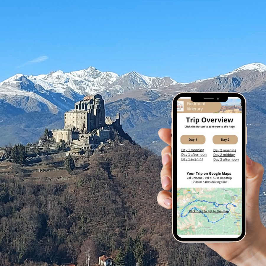 A hand holds a smartphone displaying a two-day road trip itinerary for Piedmont. In the background, the Sacra di San Michele stands against snow-capped mountains under a blue sky.