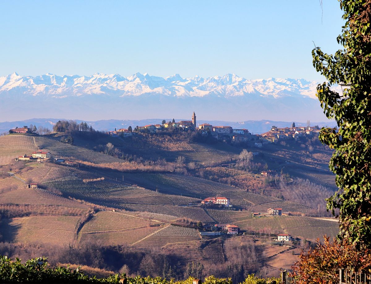 Ein Dorf thront auf einem Hügel über weitläufigen Weinbergen der Langhe, dahinter zeichnet sich die schneebedeckte Alpenkette klar am Horizont ab. Das Panorama passt ideal zu einem Langhe Roadtrip, weil es die besondere Lage der Weinregion im Piemont zwischen Rebhügeln und Alpen eindrucksvoll zeigt.