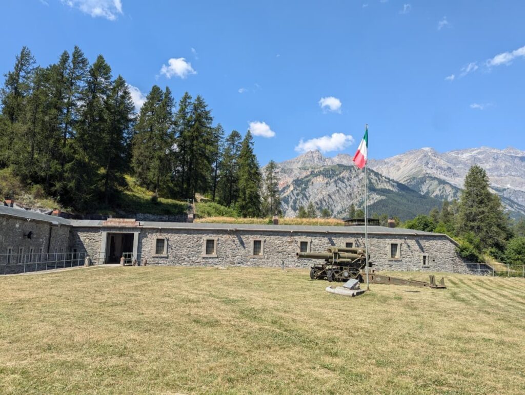 The historic Alpine fort Bramafam with an Italian flag and old artillery sits beneath the rugged mountains of Piedmont, adding a cultural stop to an itinerary for how many days to spend in Piedmont Italy.